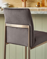 Modern bar stool with a sleek design, featuring a brushed brass metal frame and a plush, charcoal-grey upholstered seat and backrest.