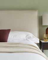 Beige upholstered headboard on a bed with white bedding, a herringbone-patterned throw at the foot, a burgundy accent pillow, a dark wooden nightstand with an open shelf below, and a white lamp with a cylindrical shade.