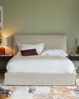 Elegant contemporary bedroom featuring a king-sized bed with a curved, upholstered beige headboard and a matching beige platform base. White bedding with an additional deep burgundy accent pillow creates a warm and inviting look. Bed is flanked by two matching dark wood nightstands with visible drawers, each hosting a round, ribbed, terracotta-coloured table lamp, casting soft light. The bed sits atop a multi-tonal area rug with abstract design in warm earth tones.