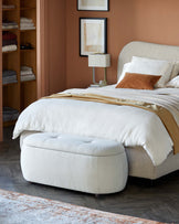 A cozy bedroom featuring a plush upholstered bed, a soft cream ottoman, and a stylish bedside lamp against a warm backdrop.