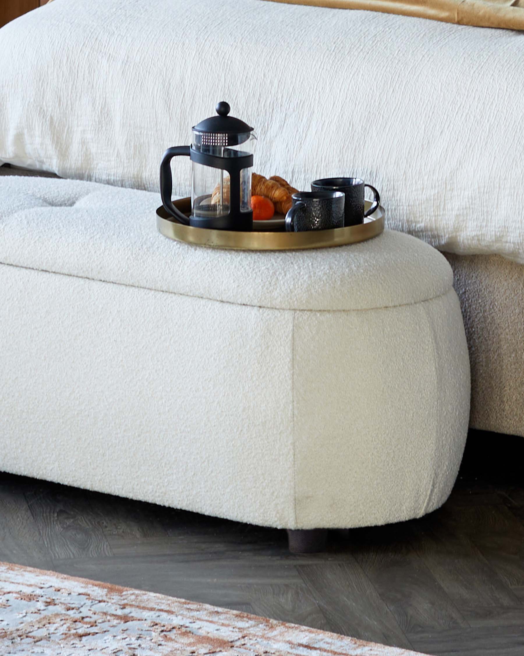 Soft, cream-colored ottoman with a round tray holding a French press, cups, and pastries, paired with a textured bed.