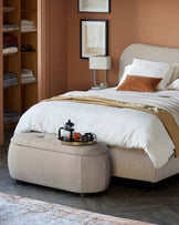 Cozy bedroom featuring a upholstered bed, textured ottoman with a tray, bedside lamp, and framed artwork against a warm wall.