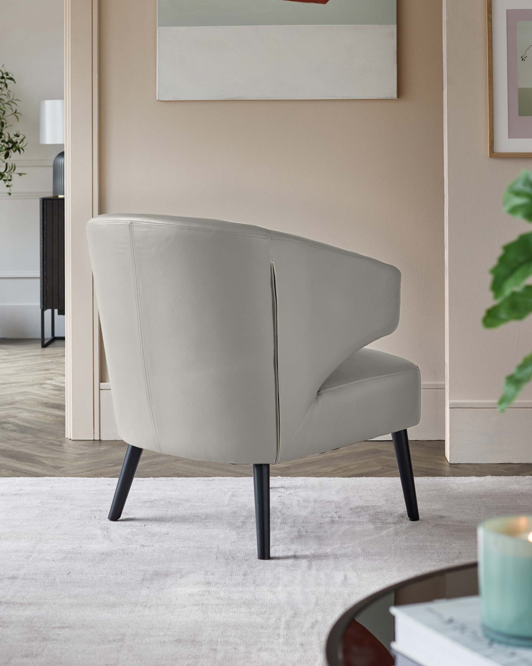 Modern light grey armchair with a curved backrest and angular design, featuring slender black legs. The chair is situated in a stylish room setting over a light-coloured area rug, with artwork and indoor plants subtly complementing the minimalist aesthetic.