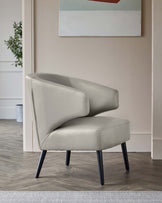 Modern light grey upholstered armchair with a sleek, curved design and four angled black legs, set in a contemporary room with wood flooring and a neutral colour scheme.