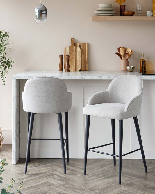 Two modern upholstered bar stools with sleek grey fabric and black metal legs are positioned at a white marble countertop. The stools feature curved backrests and integrated footrests for comfort and style.