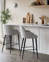 Two modern bar stools with light grey upholstery and black metal legs positioned at a kitchen island with a white marble countertop.