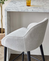 Modern dining chair with an upholstered light grey textured fabric seat and winged backrest, paired with sleek black metal legs, posed next to a marble-top table.