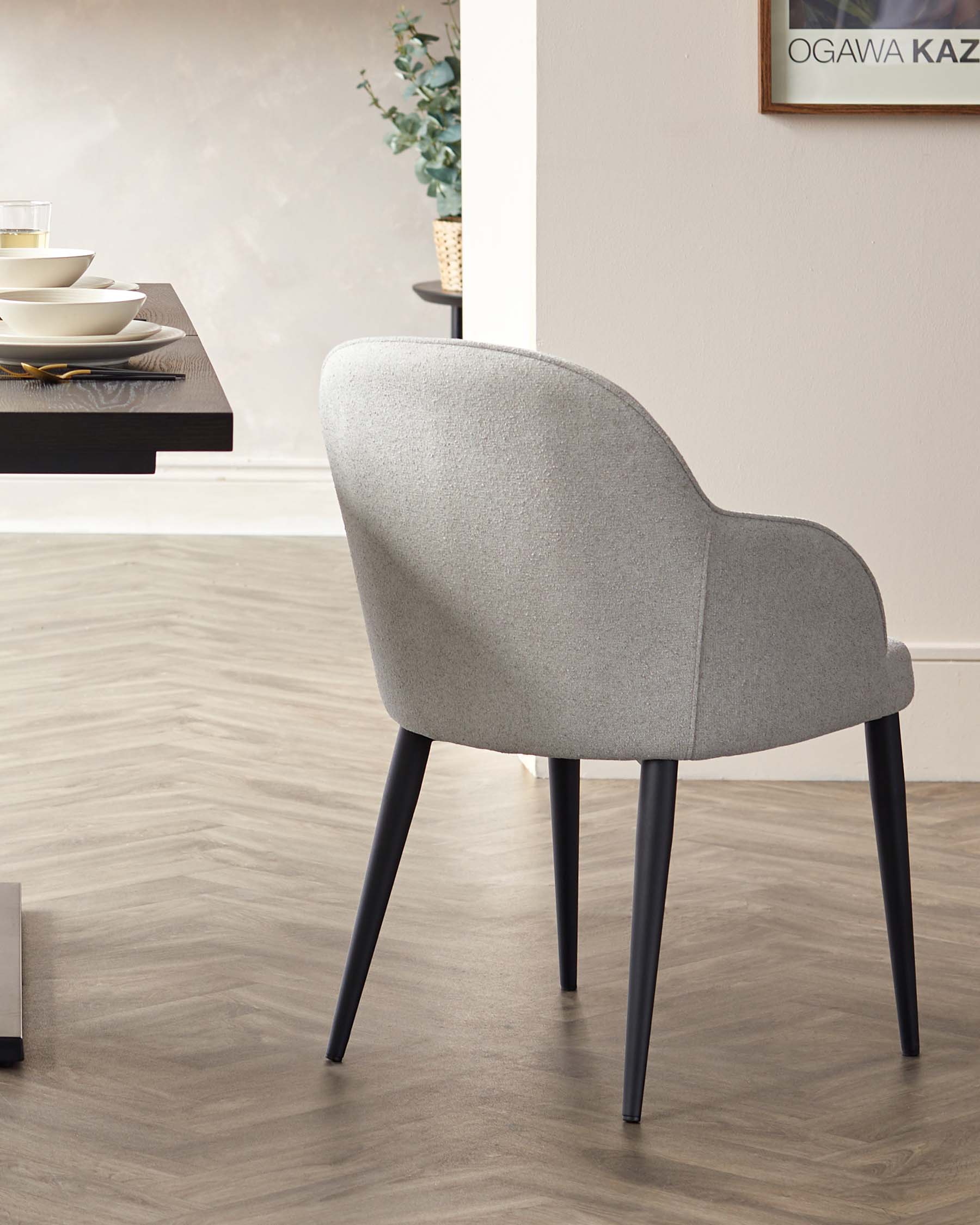 A modern light grey upholstered dining chair with a curved backrest and four sleek black legs. The chair is positioned on a hardwood floor, with a partial view of a sideboard and decorative items in the background, contributing to a minimalist aesthetic.