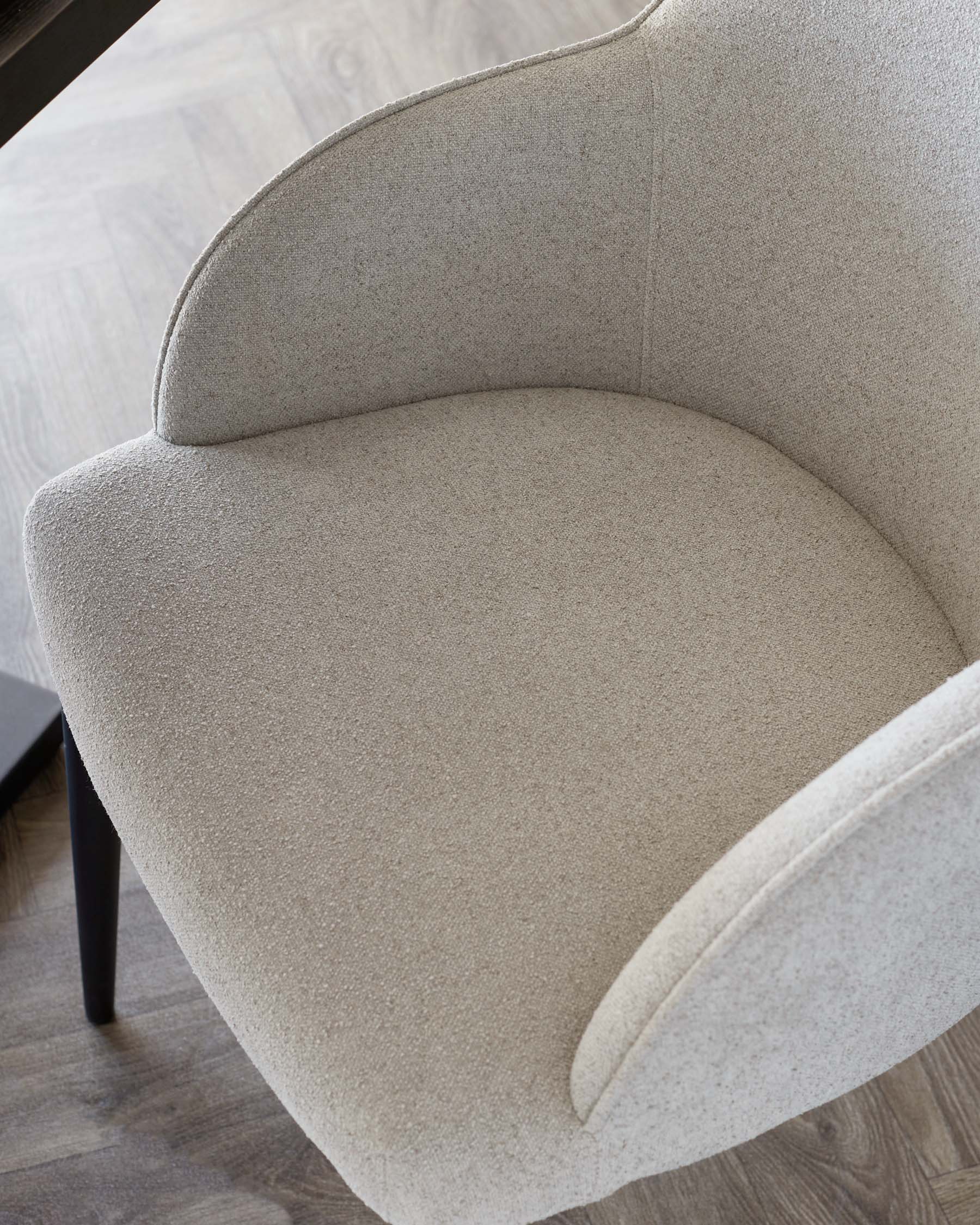 Close-up of a contemporary beige upholstered armchair with a curved backrest and dark wooden legs on a wood-patterned floor.