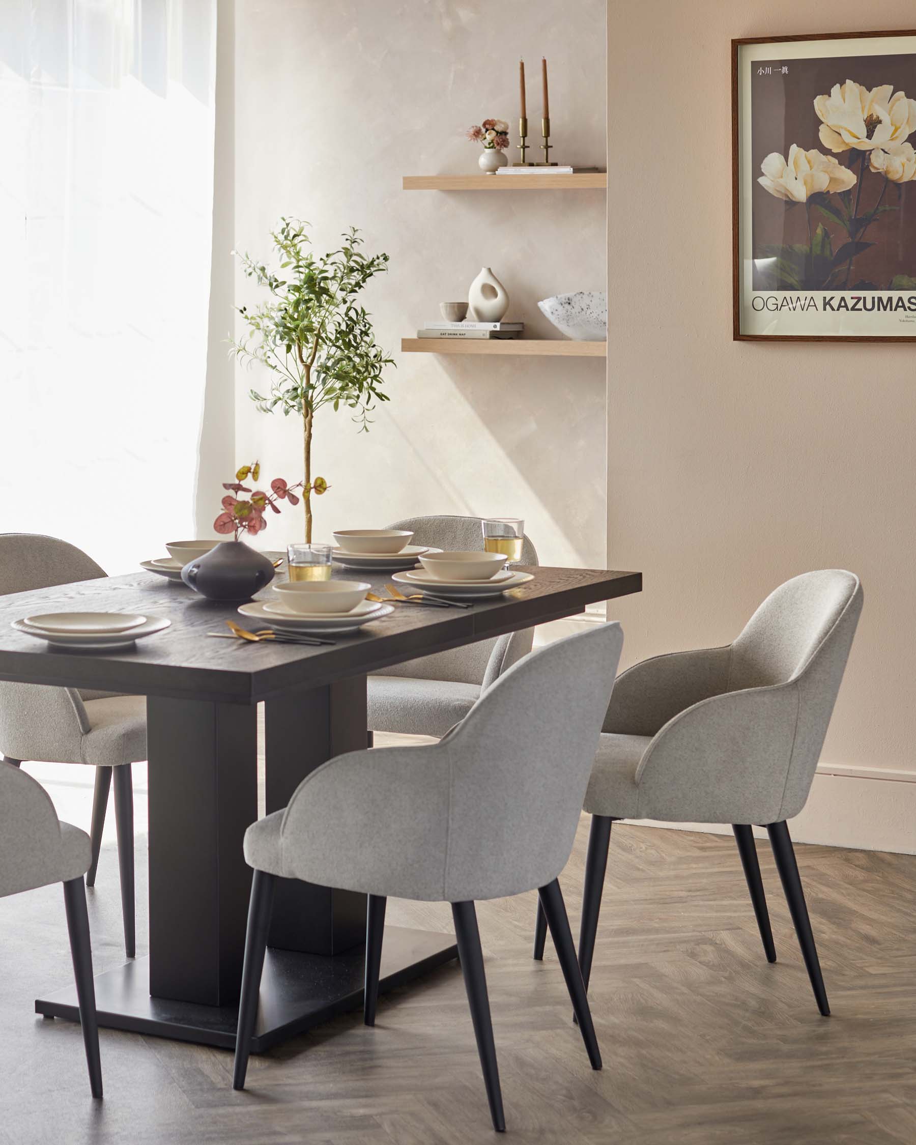 A modern dining set comprising a rectangular wooden table with a dark finish and sturdy block legs, accompanied by four upholstered chairs in a light grey fabric with a slightly curved backrest and slender black legs. Two wooden shelves mounted on the wall in the background complement the ensemble.