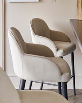 Elegant modern armchairs with light beige upholstery and slender black metal legs, positioned around a minimalist dining table with a dark wood top.