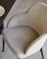 Elegant modern armchair with velvety taupe upholstery and curved backrest, featuring sleek black legs; positioned on a light wooden floor beside a portion of a dark tabletop with a striped ceramic plate visible in the top left corner.