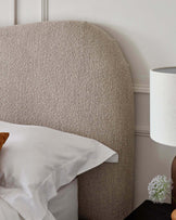 Curved upholstered headboard in a textured beige fabric, partial view of a white bedside lamp with cylindrical shade, and a glimpse of a wood nightstand with a blooming plant in a vase.