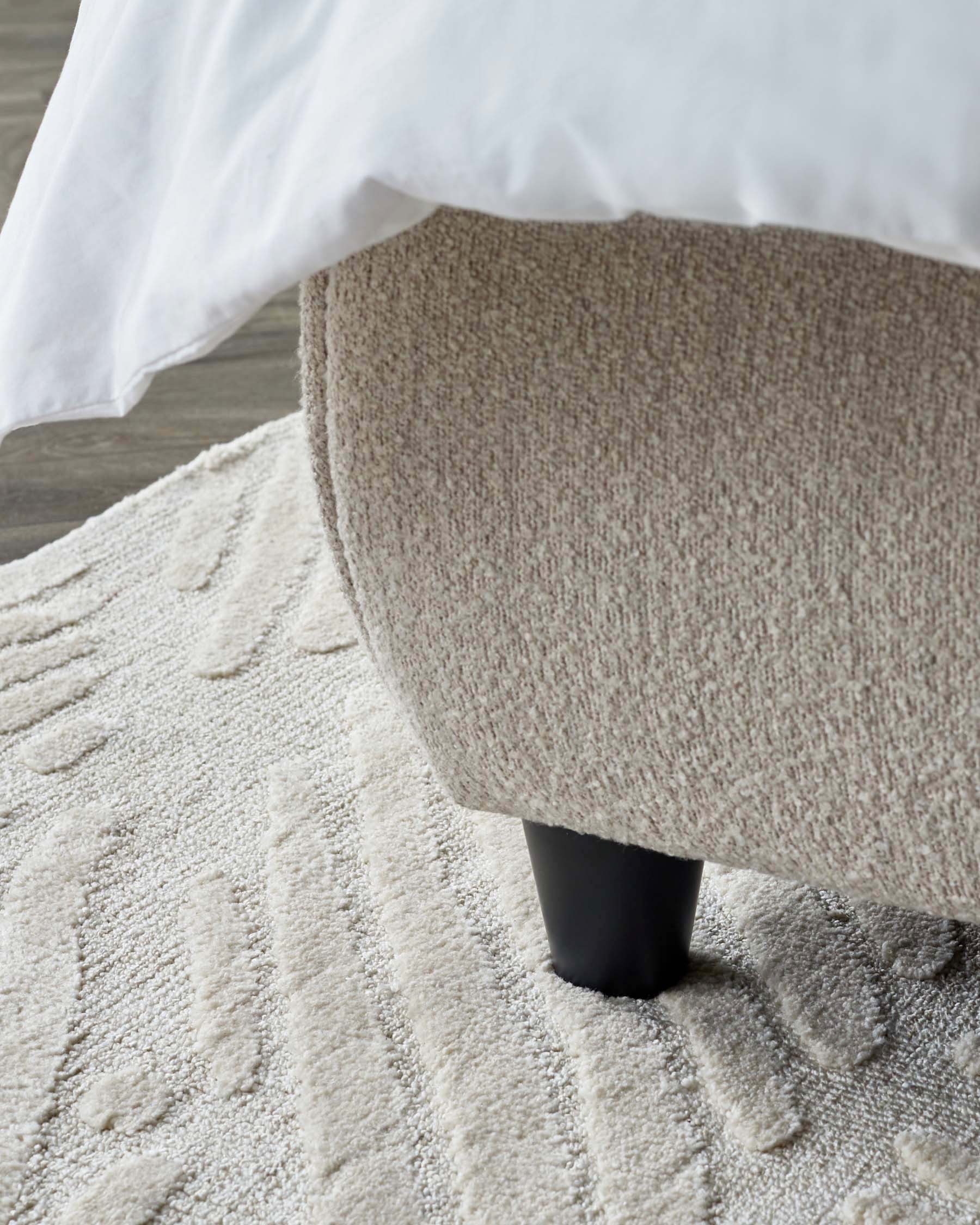 Close-up view of a piece of furniture featuring a textured beige upholstery and a dark, tapered cylindrical leg. Part of a white fabric, likely a bed sheet or cover, drapes over the corner of the piece, on top of a plush, patterned off-white rug. The flooring appears to be a grey wood or wood-like material.