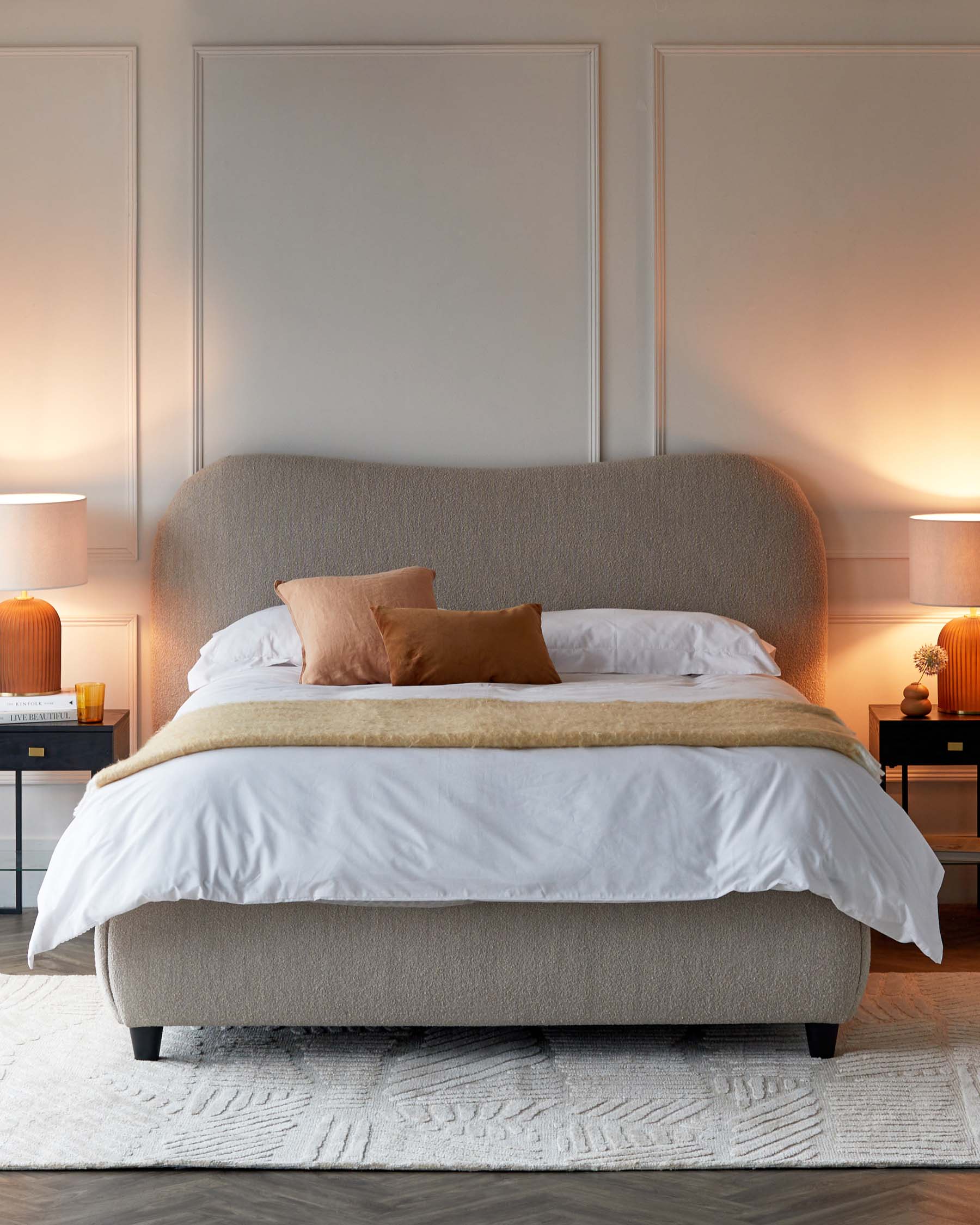 Modern minimalist bedroom setting featuring an upholstered platform bed with a curved headboard in a neutral fabric, flanking mid-century style nightstands with black frames and wood drawers, topped by matching ceramic table lamps. A textured white area rug lies beneath the bed.