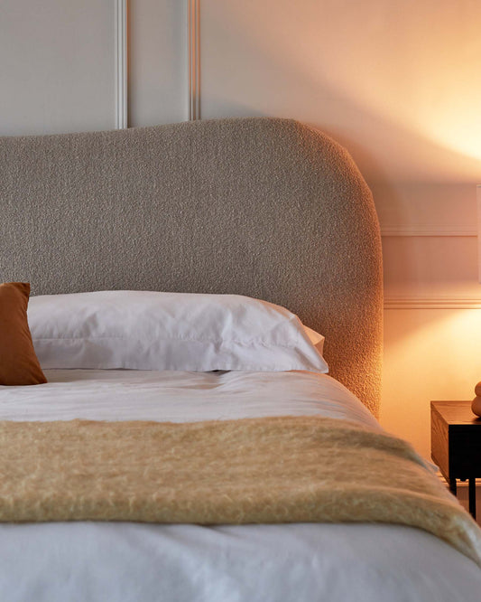 Elegant bedroom featuring a textured upholstered headboard, soft white bedding with a golden throw, and a warm-toned wooden nightstand with a table lamp.