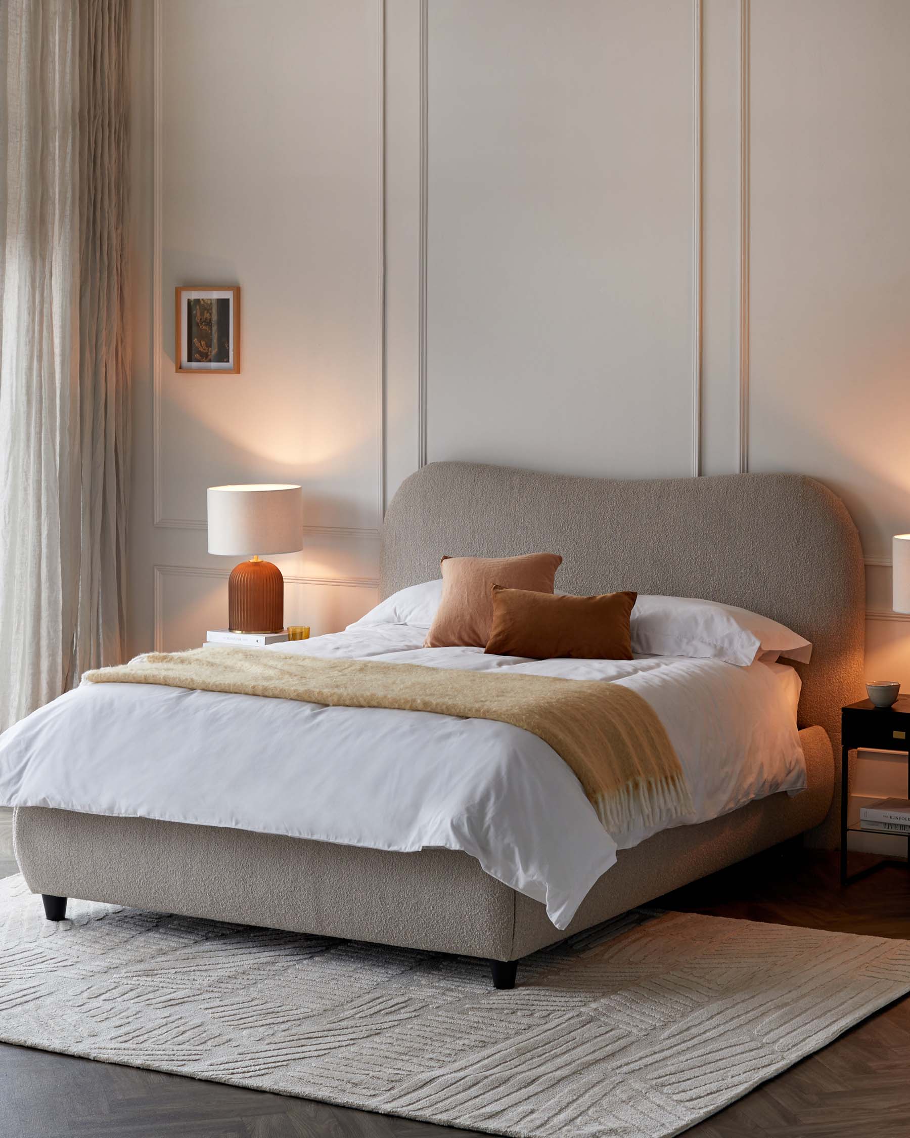 Elegant contemporary bedroom featuring a king-sized bed with an upholstered curved headboard in a light beige fabric, set on dark wooden legs. Two wooden nightstands flank the bed, one with an open shelf and the other with a drawer, both topped with cylindrical table lamps with white shades. The bed is complemented by crisp white bedding, a golden-hued throw, and matching pillows. A textured light grey area rug lies beneath the bed, adding warmth to the room's ambiance.