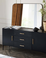 A contemporary black wood sideboard with ribbed detailing and brass handles. The sideboard is accompanied by a large, arched-top mirror with a walnut frame, resting on the surface. The scene is accessorized with a black vase, an open book, and a plant in a wicker basket, set against a background of white walls and a sheer curtained window.