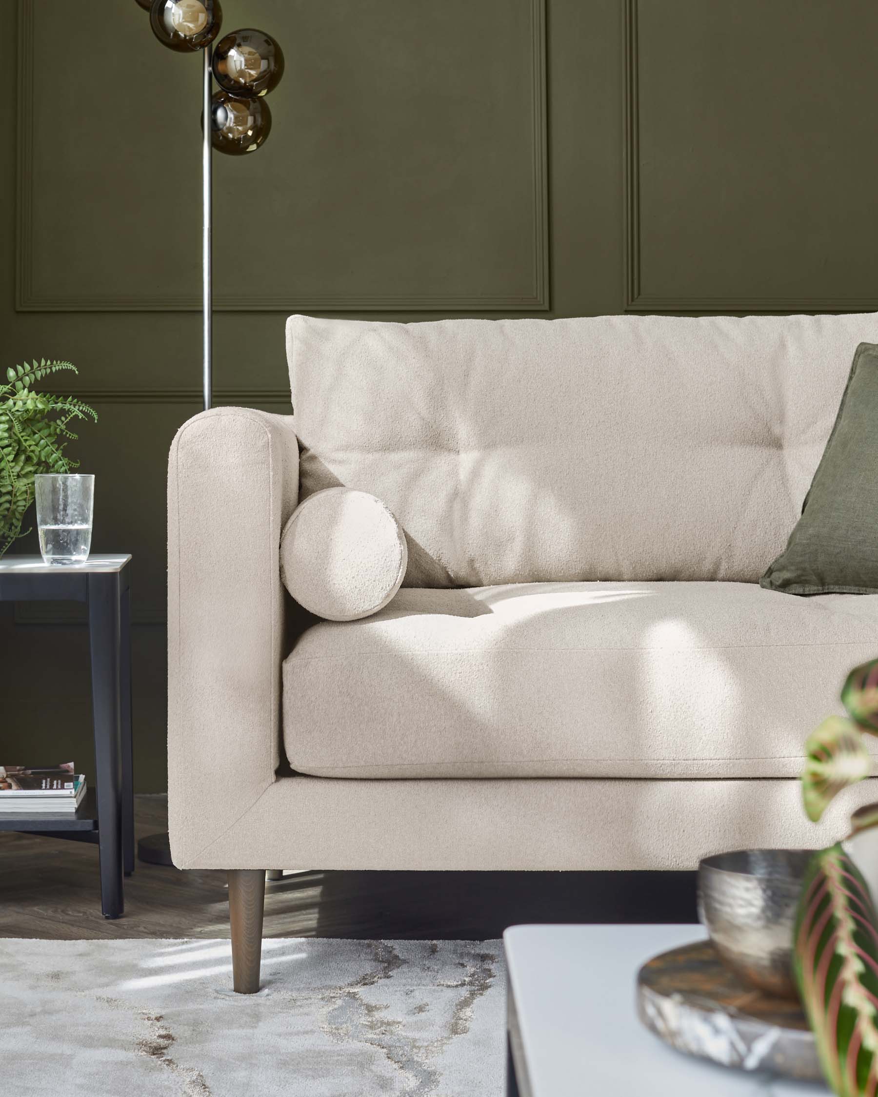 A modern beige fabric sofa with cylindrical side cushions, complemented by sleek, tapered wooden legs. A minimalist dark side table with a clear glass and a round metal bowl, a potted fern, and a stylish floor lamp with spherical shades enhance the contemporary setting. The room features a soft patterned area rug and olive green wall panels for an elegant touch.