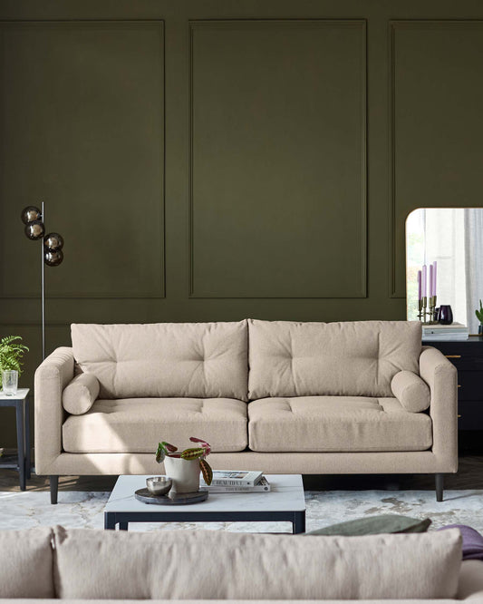 A modern beige three-seater sofa with clean lines, cylindrical cushions, and square armrests, set in a living room. A mid-century modern style floor lamp with spherical shades stands to the left. A rectangular grey coffee table with a white top displaying decorative items and books is in the foreground. Behind the sofa, a black sideboard with tapered legs and dark purple candles on top completes the scene, all against a wall with green panelling.