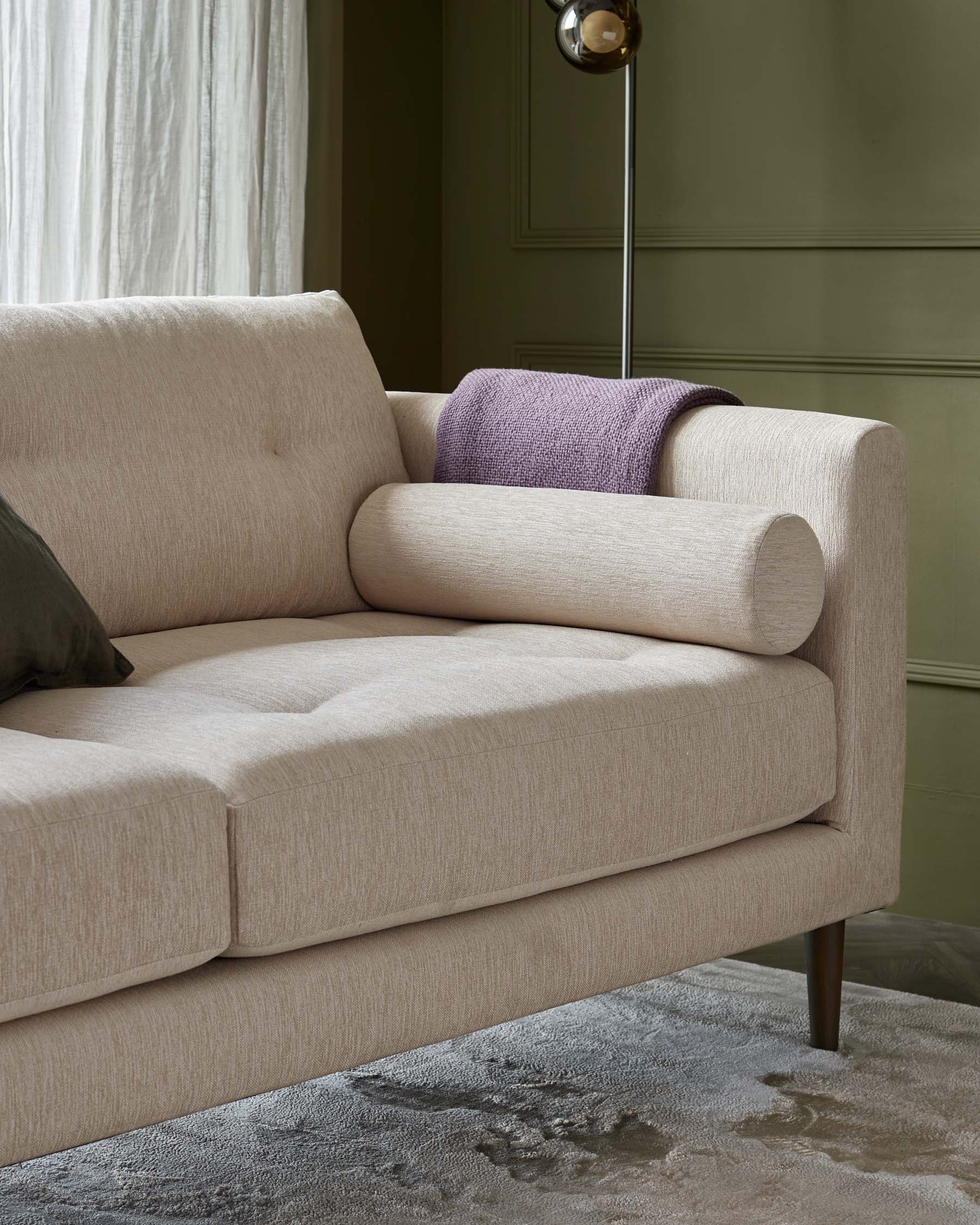 Beige fabric upholstered sofa with cylindrical bolster pillows, featuring splayed wooden legs, and set against an olive green wall with a cream curtain and brass-coloured floor lamp peeking from behind. A purple textured throw pillow adds a pop of colour. The sofa rests on a textured grey area rug.