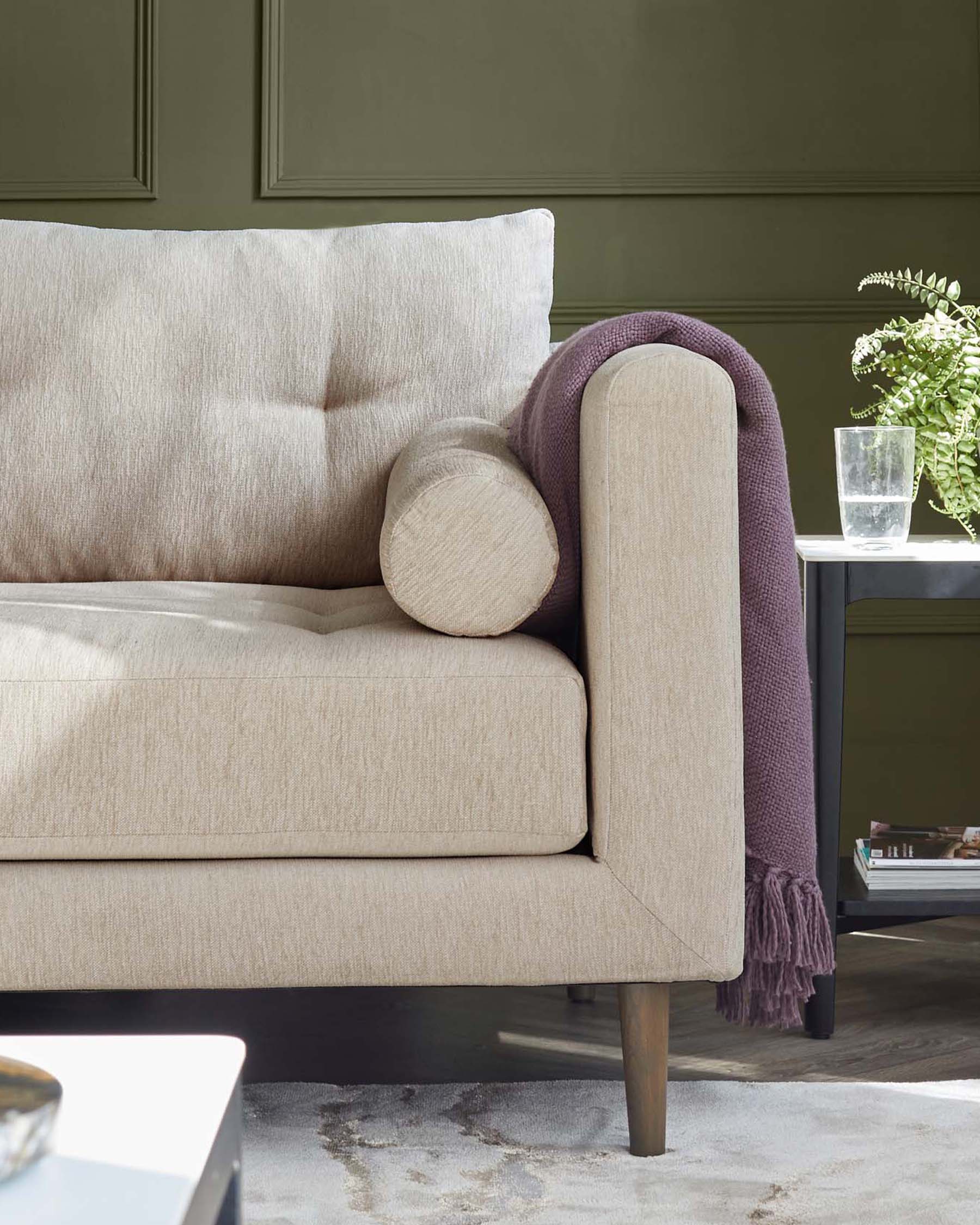 Elegant beige fabric sofa with cylindrical bolster pillow, featuring clean lines, cushioned back and seat, dark wooden tapered legs, accessorized with a textured deep purple throw blanket draped over the side. A small round side table with a glass top and a greyscale mat beneath are also visible, showcasing a clear glass of water and a decorative potted fern, completing the cosy, refined setting.