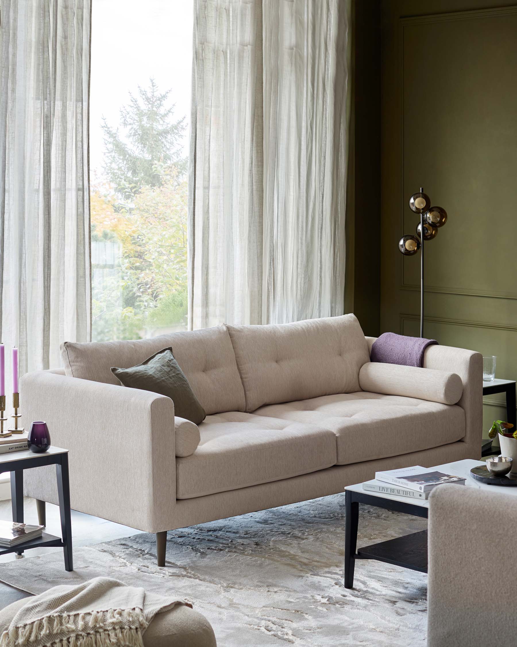 Modern minimalist living room with a beige upholstered three-seater sofa with clean lines and plush cushions. Near the sofa is a squared black side table with a lower shelf displaying a small pile of books. In the foreground, part of a matching beige armchair can be seen, and a low coffee table with books and decorative items rests on a textured grey area rug. Elegant sheer curtains frame a window providing natural light, enhancing the room's serene and sophisticated ambiance.