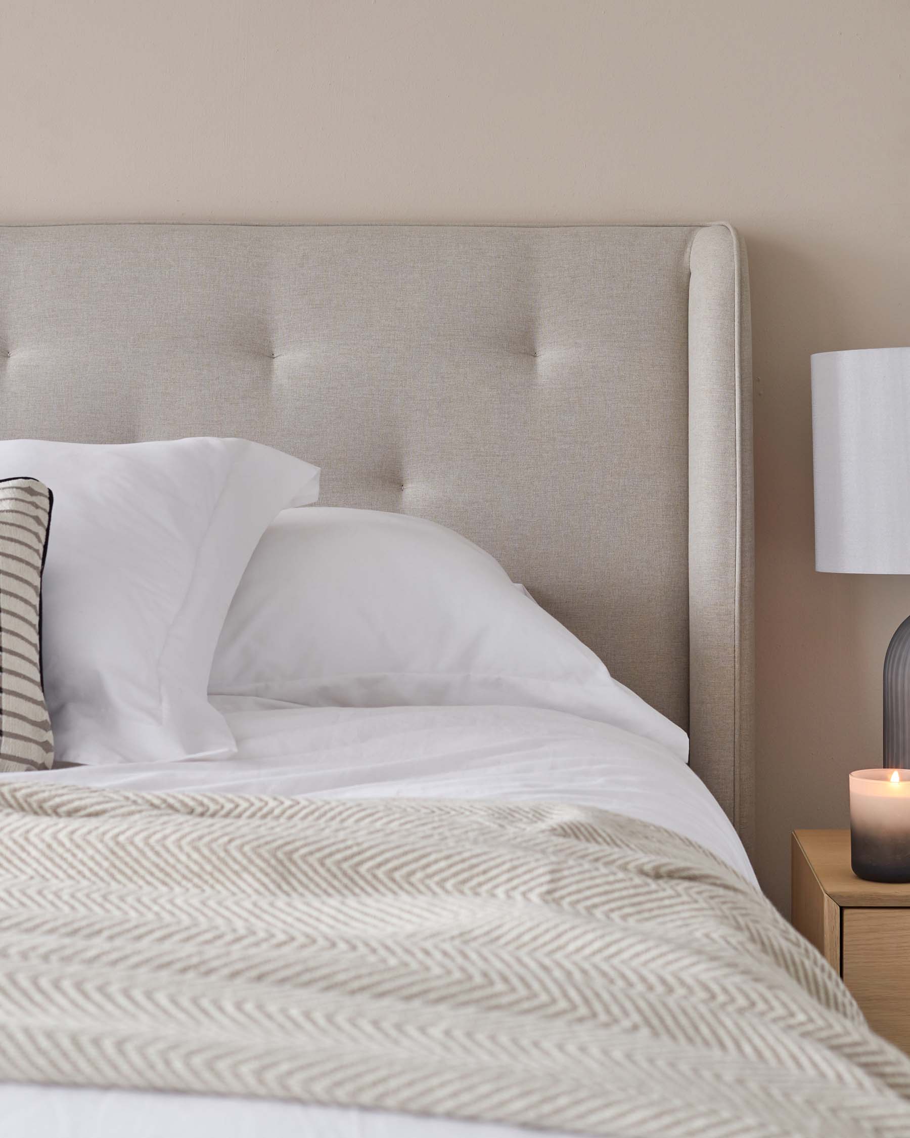 A modern upholstered headboard in a neutral fabric with tufting details, paired with crisp white bedding with a subtle herringbone pattern. A sleek bedside table with a simple drawer and a contemporary table lamp with a white shade sits beside the bed. A small candle provides a touch of ambiance.