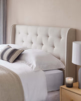 Elegant bedroom scene featuring a tall, tufted beige headboard with a softly curved top edge, flanking a bed dressed in crisp white linens. Beside the bed is a light wood nightstand with a simple drawer and brass handle, atop which sits a contemporary white table lamp with a cylindrical shade, next to a lit candle in a glass holder.