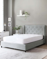 Elegant bedroom scene featuring a contemporary light grey upholstered bed frame with tufted headboard, a white two-drawer nightstand with tapered legs, and a minimalist white floating shelf above the bed. A neutral-toned rug anchors the space.