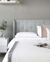 Elegant modern bedroom featuring a large upholstered headboard with button-tuft detailing in a subtle grey fabric. The bed is adorned with crisp white bedding accented with a patterned throw blanket. Beside the bed stands a contemporary white nightstand with a textured front, subtly adorned with minimalistic decor and a single green potted plant, contributing to a serene and sophisticated atmosphere.