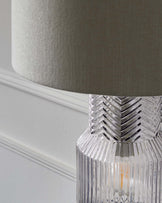 Close-up of a contemporary table lamp with a textured clear glass base and a neutral-toned drum lampshade.