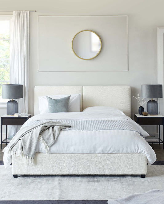 Elegant bedroom featuring a large, upholstered platform bed with a white textured headboard and footboard, flanked by two black metal end tables with square tops. Each end table is adorned with a matching dark lamp with a sizable round shade. The room also includes a simple, round gold-rimmed mirror on the wall above the bed.