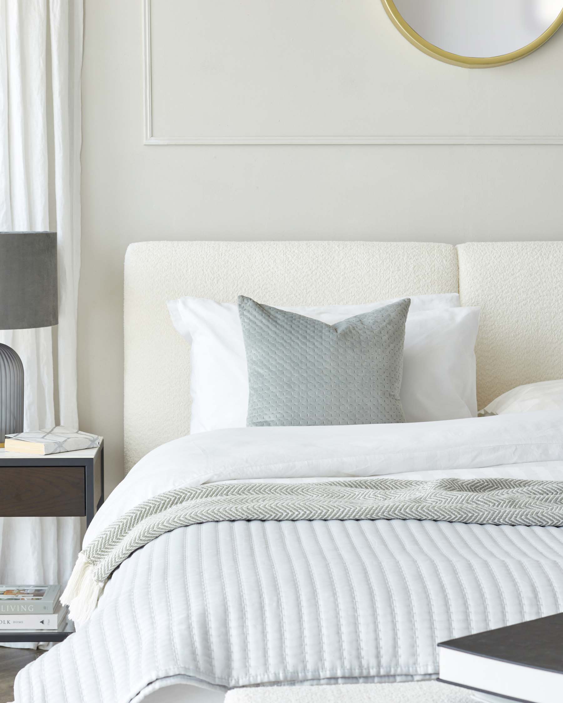 A contemporary bedroom scene featuring a cream upholstered headboard and a sleek, black bedside table with an open shelf and a drawer. A round, gold-framed mirror hangs above the bed.