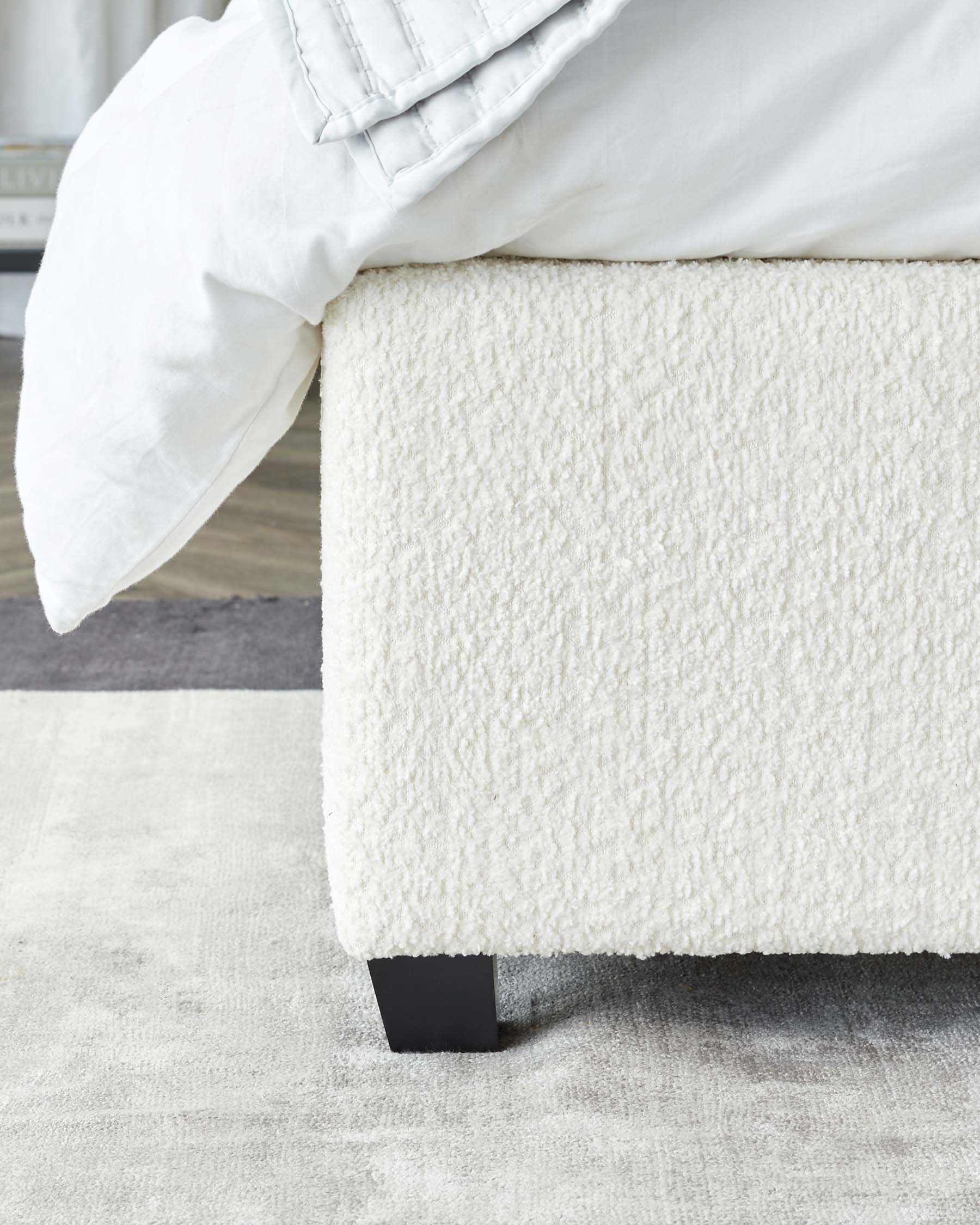 Close-up of a contemporary bed's lower corner with plush white upholstery and a dark wooden leg on a light grey rug. A white duvet and pillow partially drape over the side.