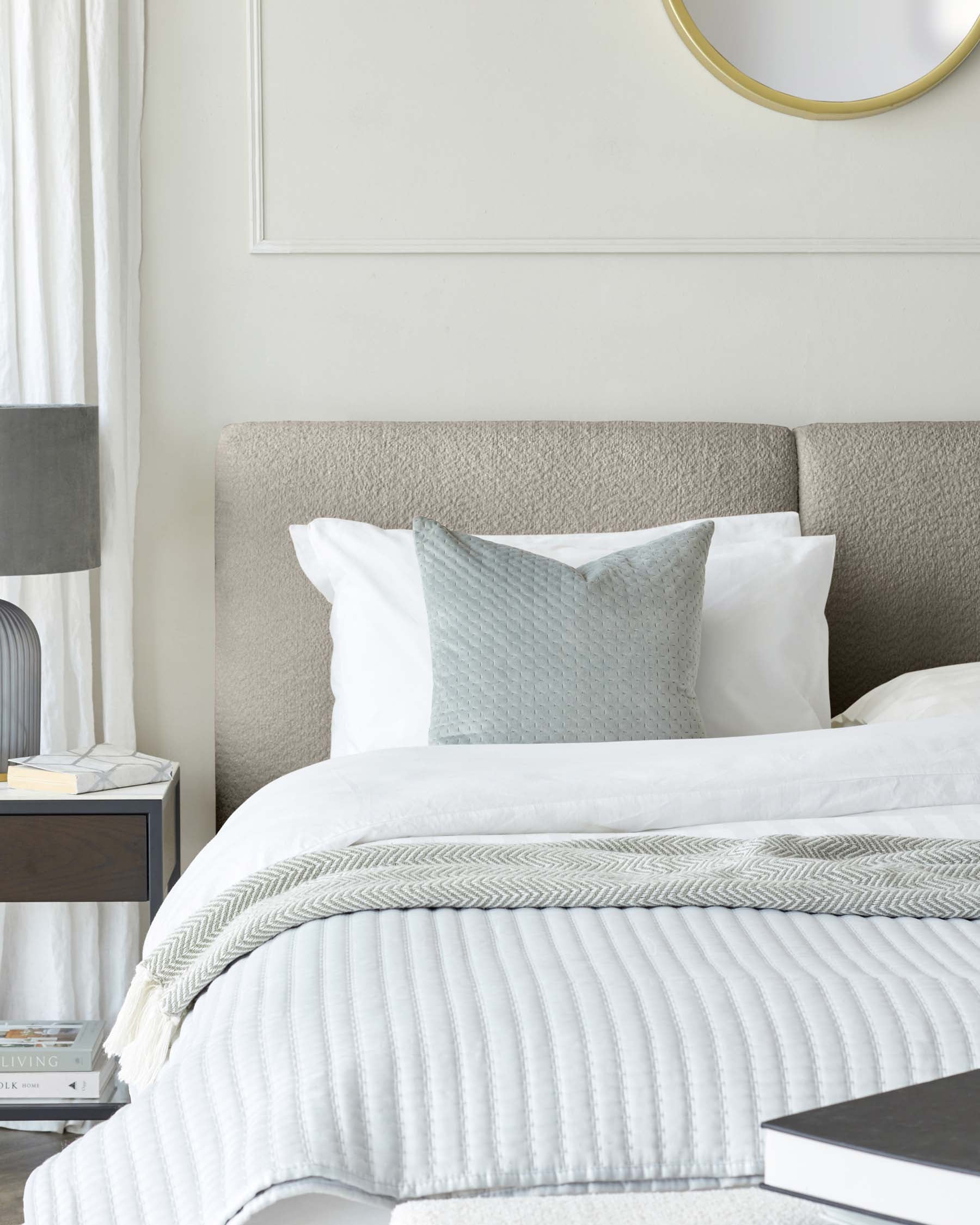 Modern bedroom furniture including an upholstered headboard in a textured neutral fabric, a minimalist black bedside table with a grey table lamp, and a white textured bedspread with decorative pillows. A circular gold-rimmed mirror hangs on the wall above the bed.