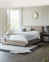 Modern minimalist bedroom featuring a large upholstered platform bed in a neutral beige tone with a textured headboard, crisp white bedding, and assorted grey accent pillows. A contemporary nightstand with a dark finish sits beside the bed, topped with a sleek table lamp and decorative items. A layered area rug combination under the bed adds depth to the room's design.