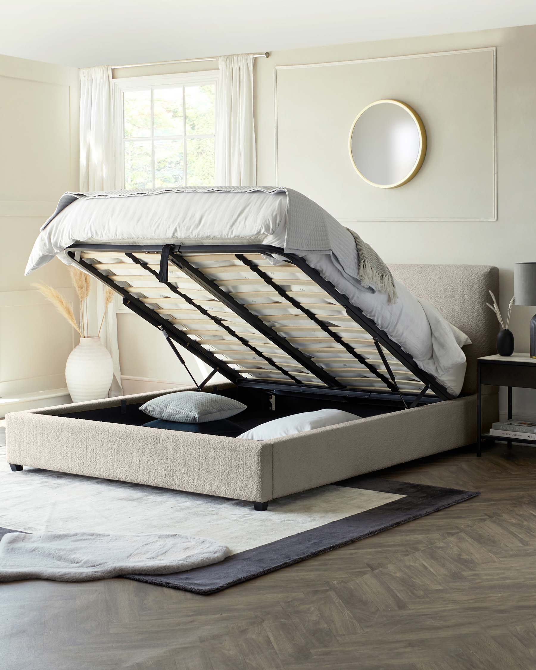 Contemporary upholstered storage bed with a lifted mattress base revealing a spacious storage area, set in a neutrally-toned room with wooden flooring and a circular wall mirror.