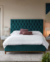 Elegant bedroom featuring a teal upholstered platform bed with a tall, tufted headboard, flanked by two white mid-century modern style nightstands with drawers and contemporary table lamps with cylindrical shades. The bed is dressed in crisp white linens and accented with a burgundy throw pillow. A large textured rug under the bed adds warmth to the space.