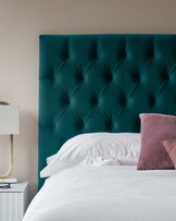 Elegant teal upholstered headboard with button tufted detailing, white bedding on a bed, and a minimalist white bedside table featuring a modern lamp with a white shade.