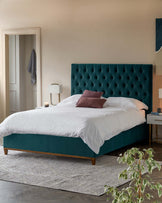 A contemporary bedroom scene with a plush king-sized bed featuring an oversized, tufted teal headboard with a diamond pattern. The bed is dressed with crisp white linens and accented with a burgundy throw pillow. A stylish white and brass nightstand with a sleek design is placed beside the bed, supporting a minimalist lamp with a white shade and a brass base. A textured grey area rug lies under the bed, adding warmth to the wooden flooring.