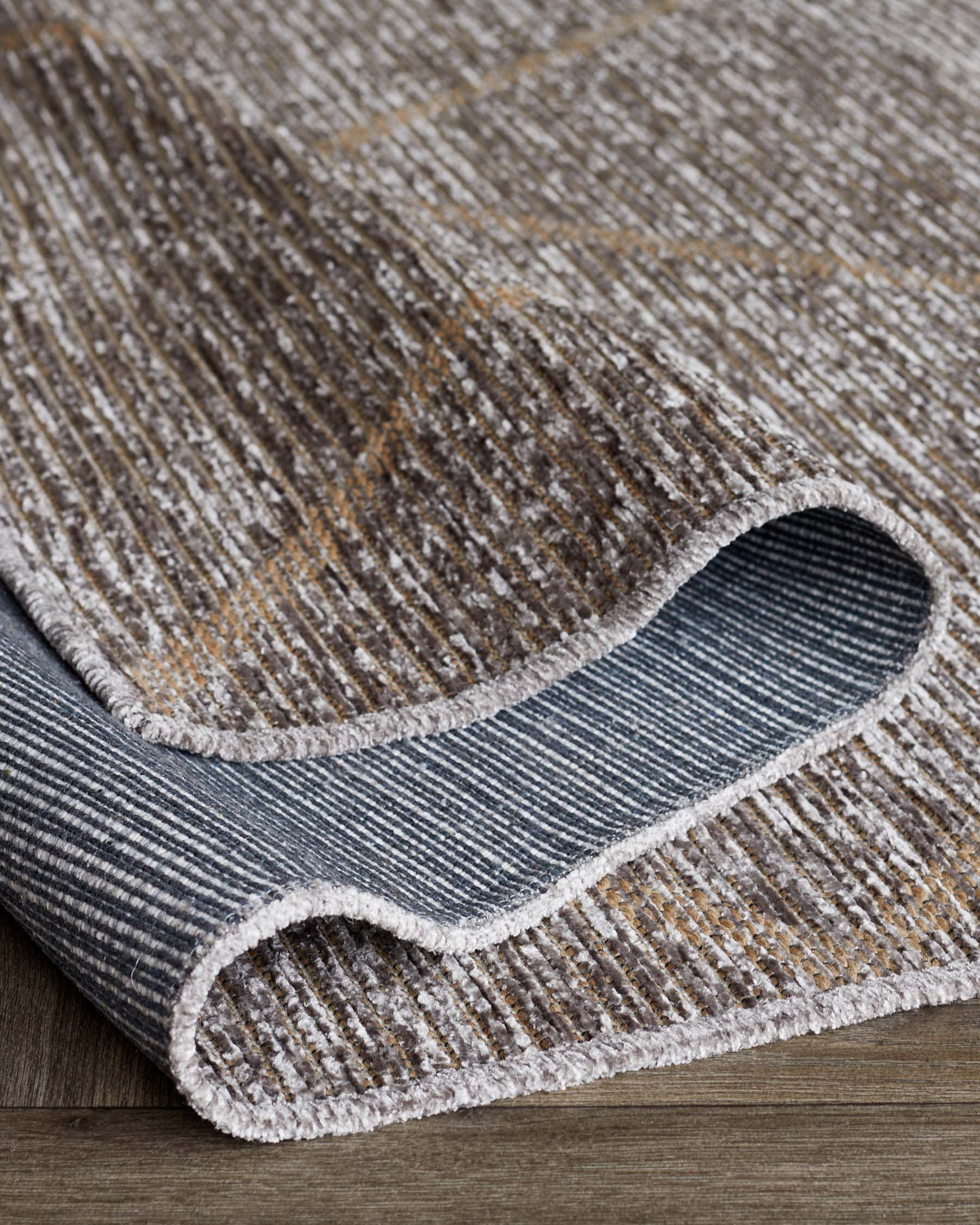 Luxurious textured area rug with a contemporary design, featuring shades of grey and hints of warm brown tones with a contrasting white edge binding, partially rolled to showcase the pattern and thickness.