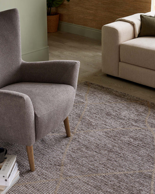 Modern living room set featuring a minimalist grey upholstered armchair with sleek lines and wooden legs, paired with a neutral-toned, contemporary fabric sofa highlighted by clean design and comfortable cushions. A patterned area rug in shades of grey and beige underlines the seating arrangement.