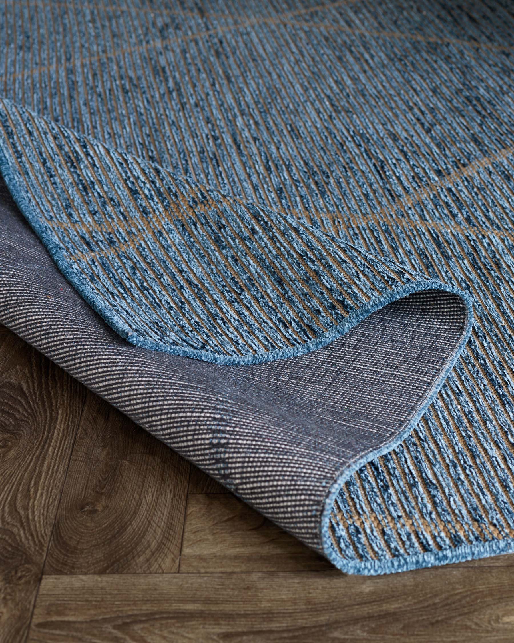 A textured blue and grey area rug with a diagonal rib pattern, partially rolled up to show its weave and underside, laid out on a wooden floor.