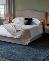 Elegant bedroom furniture arrangement featuring a king-sized bed with an upholstered headboard in a neutral fabric, crisp white bedding, a textured throw blanket with fringe detailing, complemented by a pair of modern bedside tables in a white finish, and situated on a large blue woven area rug.