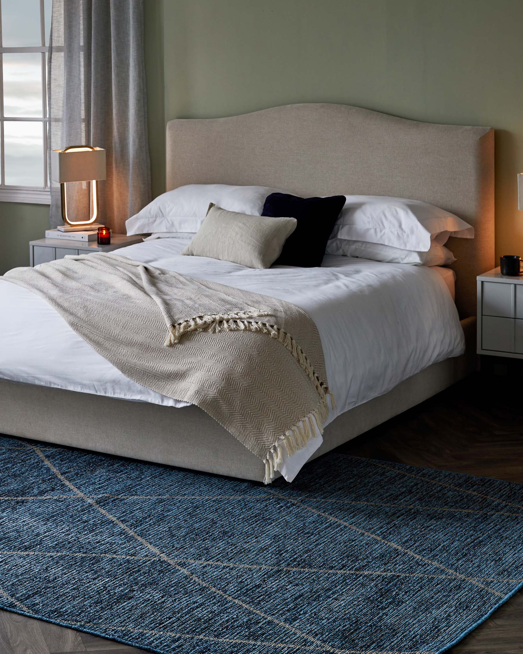 Elegant bedroom furniture display featuring a large, curved headboard upholstered in a neutral beige fabric attached to a matching platform bed frame, also upholstered. Beside the bed, there is a small, contemporary white nightstand with sleek lines. The scene is completed with a deep blue woven area rug laid out on the floor, adding a touch of colour and texture to the room.