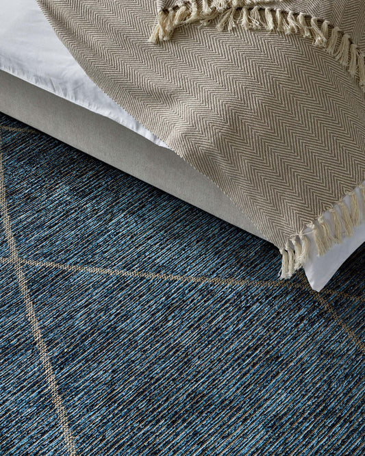 Part of a modern, minimalist platform bed with a grey upholstered frame and a textured throw blanket with fringe detailing partially covering a crisp white bed sheet, placed on top of a contemporary dark blue area rug with a geometric stripe pattern.