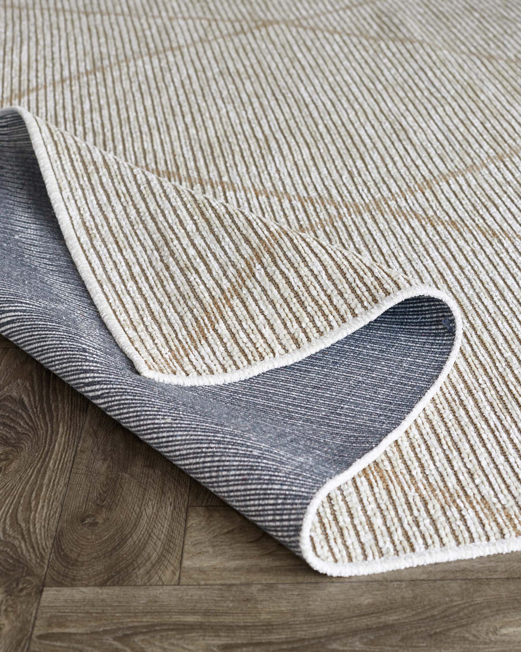 A contemporary beige and grey striped area rug with a corner rolled up, revealing a non-slip backing, laid on a wooden floor.