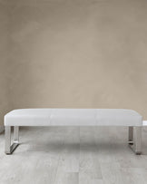 A modern white leather upholstered bench with sleek metallic legs situated on a light wooden floor against a neutral wall.