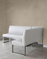 Modern white leather sofa with a minimalist design featuring sleek, chrome-finished metal legs and a lightly padded, streamlined form, set against a neutral wall on a light wooden floor.